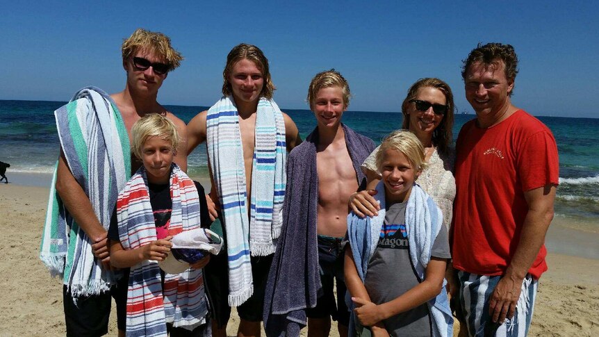 A family including mum and dad and five boys of various ages stands posing for a photo with towels on a beach.