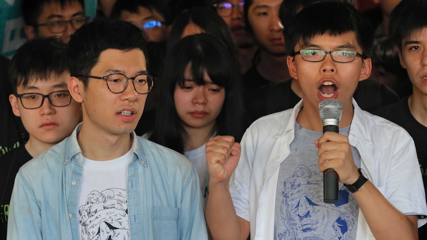 Hong Kong activists Joshua Wong and Nathan Law. Wong is speaking into a microphone. They are surrounded by young supporters.