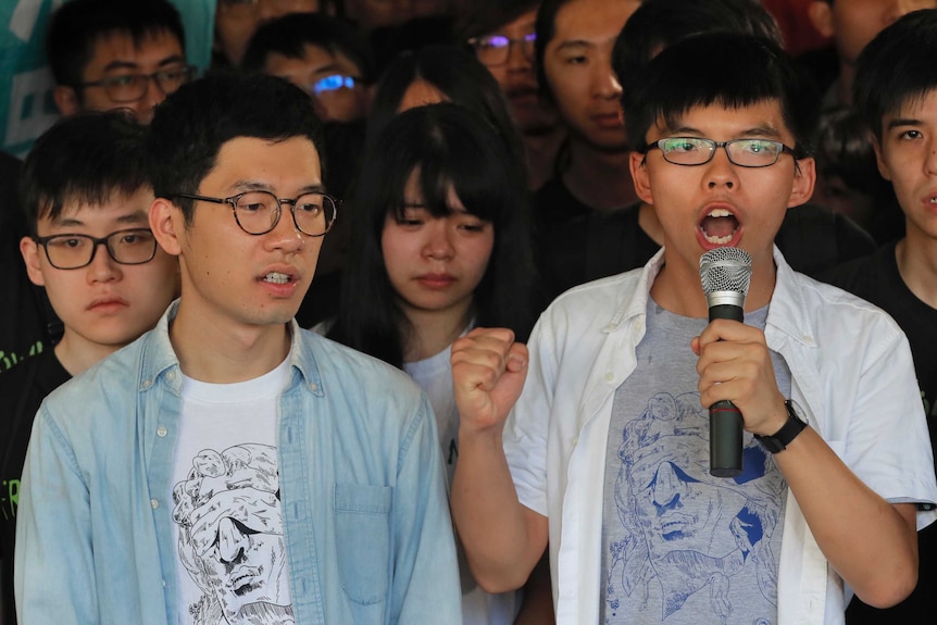 Hong Kong activists Joshua Wong and Nathan Law. Wong is speaking into a microphone. They are surrounded by young supporters.