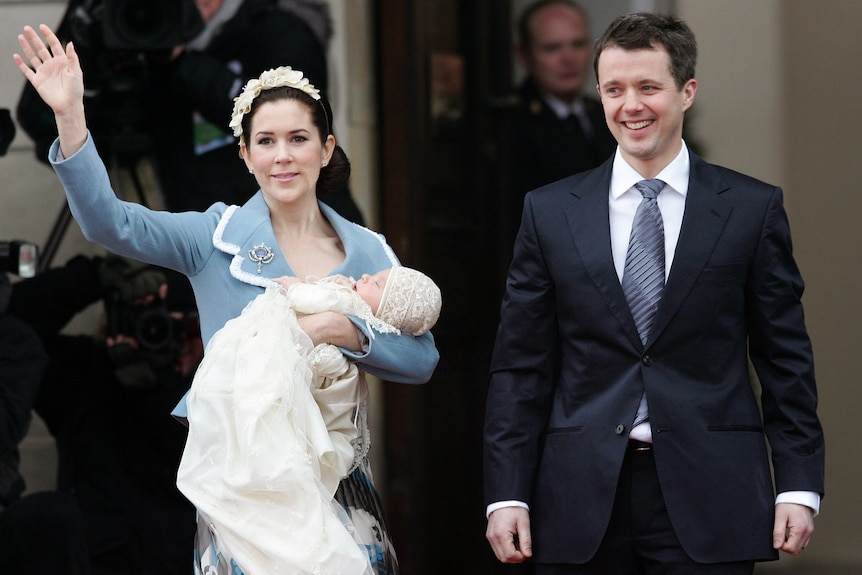 A woman in a blue suit waves while holding a baby next to a man in a suit 