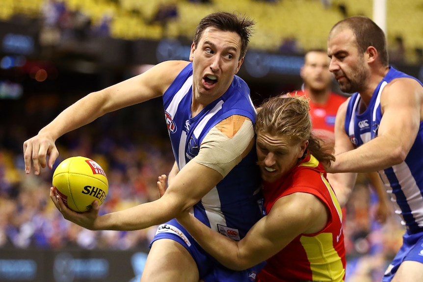 Sam Wright holds a yellow AFL ball in one hand and holds the other hand near it as a man in a red jumper tackles him
