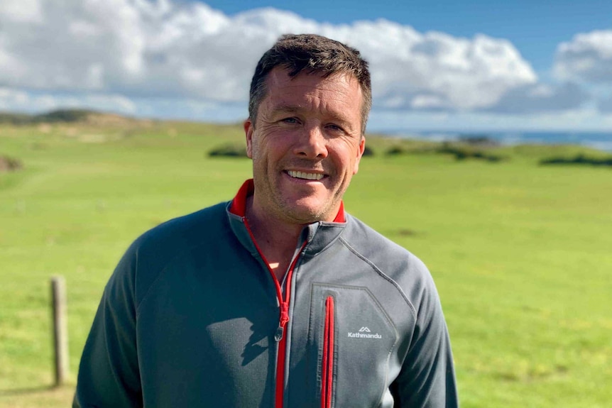 A man stands in a field on King Island, the sea is visible in the distance in the background