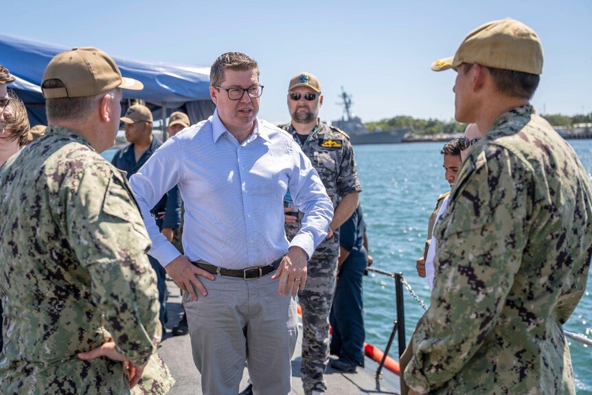 Conroy stands with hands on hips, surrounded by men in military fatigues, on board a ship deck, ocean waters in the background.