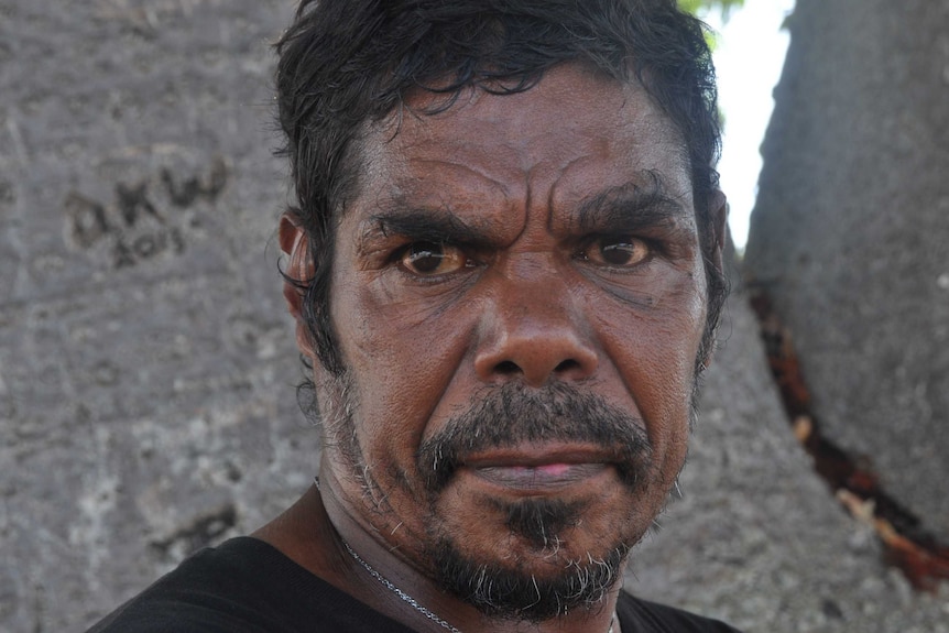 An Aboriginal man looking upset standing in front of a tree.