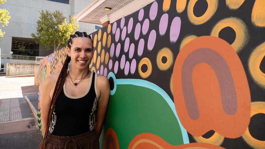 A smiling young woman with a long corn-row hairstyle, stands beside a colourful Indigenous art mural.