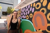 A smiling young woman with a long corn-row hairstyle, stands beside a colourful Indigenous art mural.