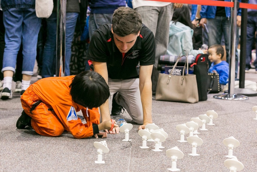 A man kneels on the ground with a child and small models of a telescope.