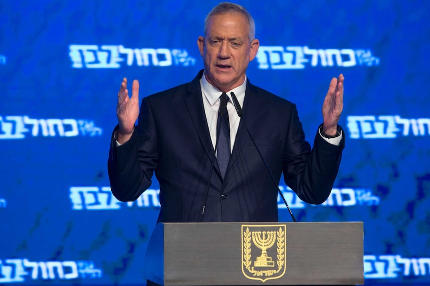 Benny Gantz wears a blue suit and speaks at a podium in front of a bright blue party poster