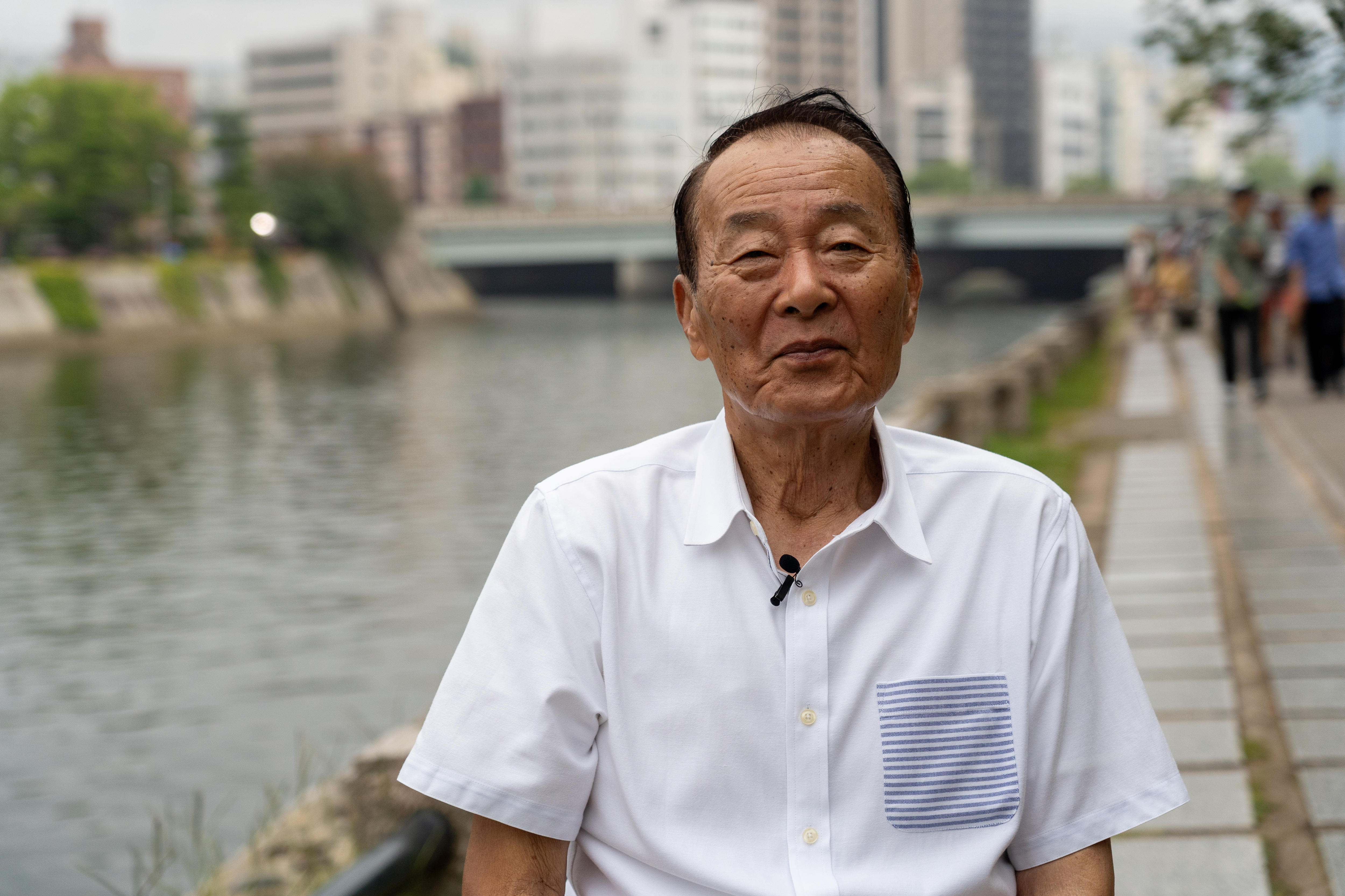 A Japanese man wearing a white button-up collar t-shirt standing on a tiled footpath next to a waterway