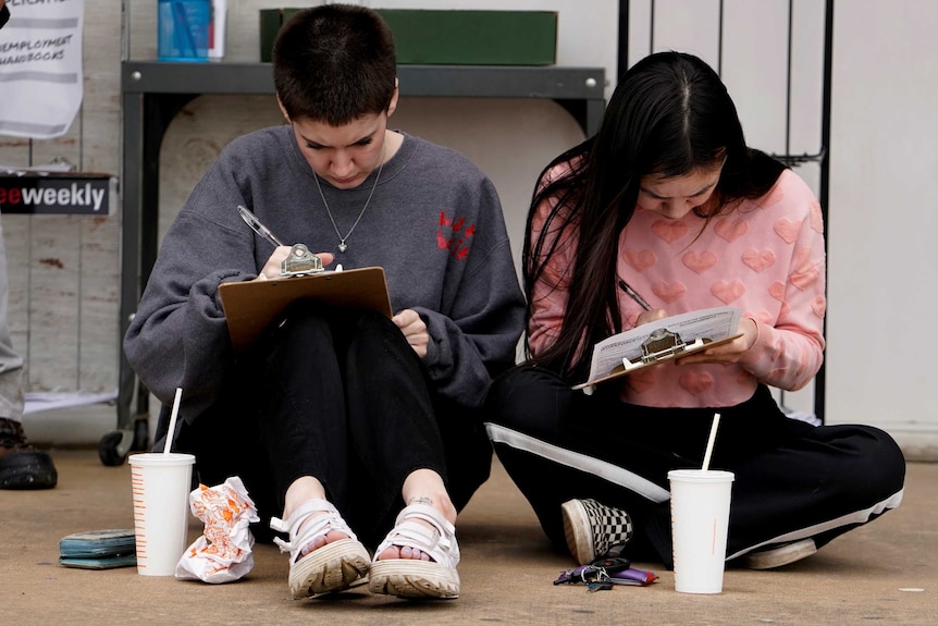Two young women sitting on the ground filling out unemployment papers on clipboards