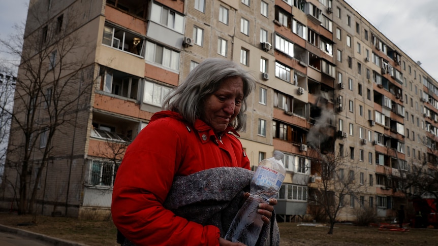 A woman clutches a water bottle as her burntout apartment building is seen behind her. 