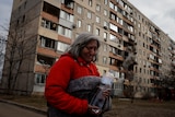 A woman clutches a water bottle as her burntout apartment building is seen behind her. 
