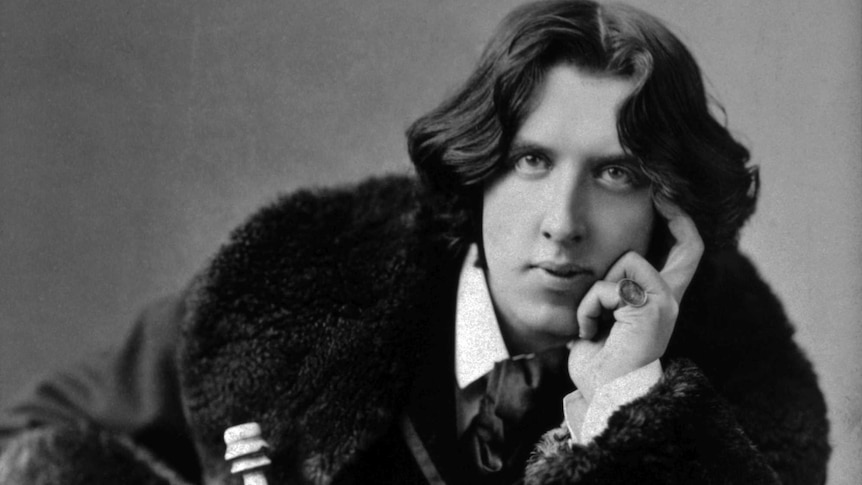 black and white portrait of seated man with wavy dark hair leaning forward, elbow on leg