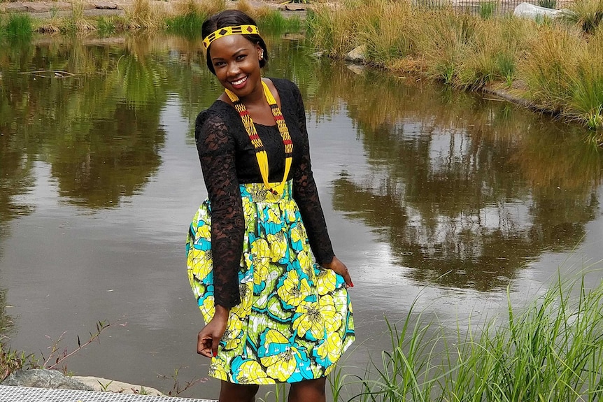 A woman stands in front of a pond wearing traditional South Sudanese dress.