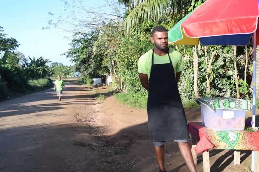 Gideon Rambe sells homemade donuts, known as gateau, by a dirt road. He has a colourful rainbow umbrella over his stall.