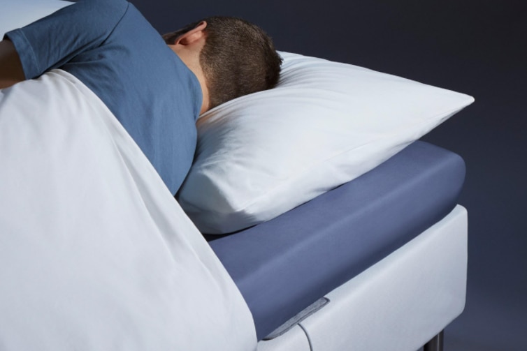 A brown haired man wearing a blue top sleeps on his side facing away. There is an edge of a grey mat underneath the mattress