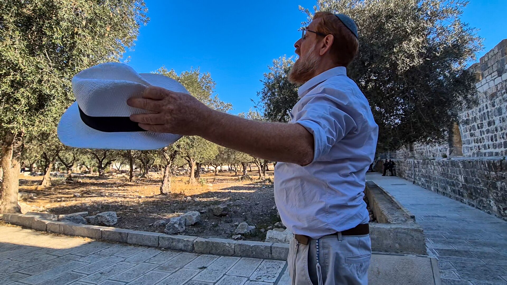Yehuda Glick stands on a stone path with his arms raised at his sides, with a hat held in his left hand.