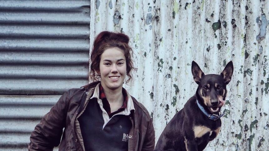 a photo of a young woman in jeans and farm wear with a kelpie smiling at the camera