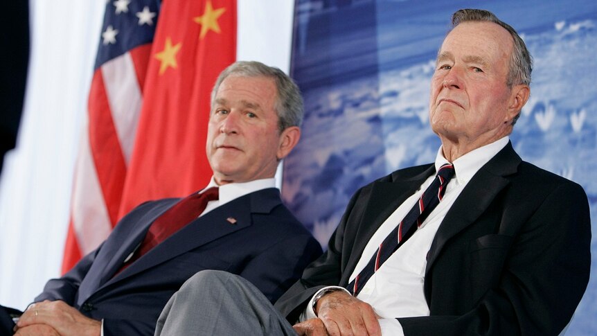George W Bush and his father George HW Bush look concerned as they sit in front of an American flag.