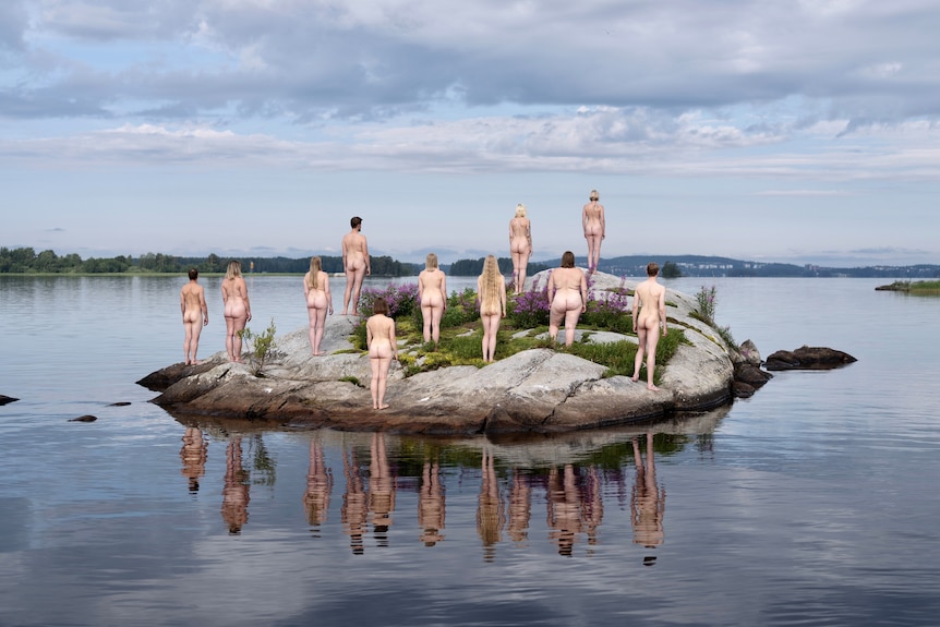 A photograph of 11 naked people standing on a rock, surrounded by water, facing away from the camera