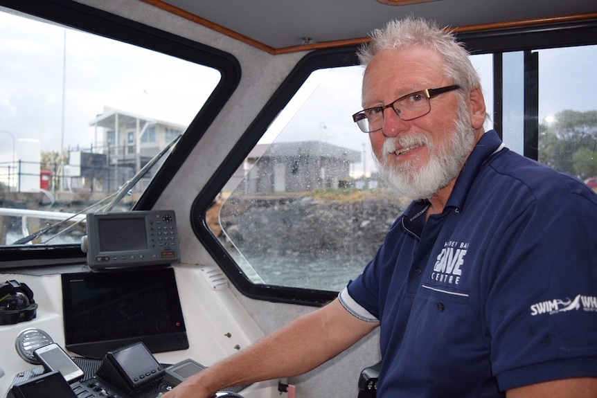 A man with grey hair, grey beard and glasses, wearing a blue polo shirt, sits at the controls of a boat.