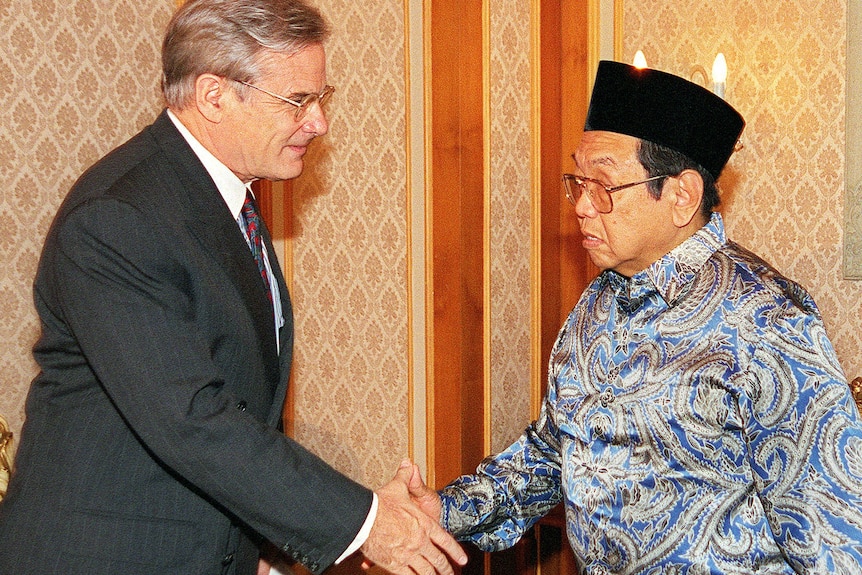John McCarthy, in a dark suit, with grey hair and glasses, smiles as he shakes hands with Abdurrahman Wahid