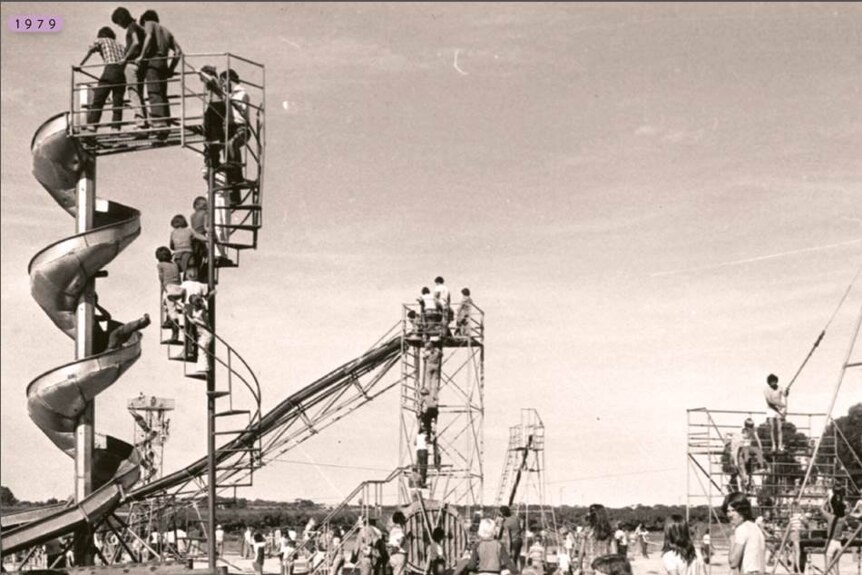 A black and white photograph showing a playground in 1979 with lots of people on rides including a 15 metre high spiral slide.