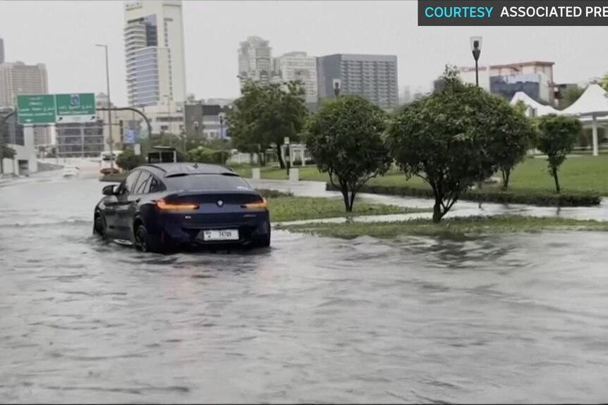 Cars drive through a flooded roadway with cityscape in the background.