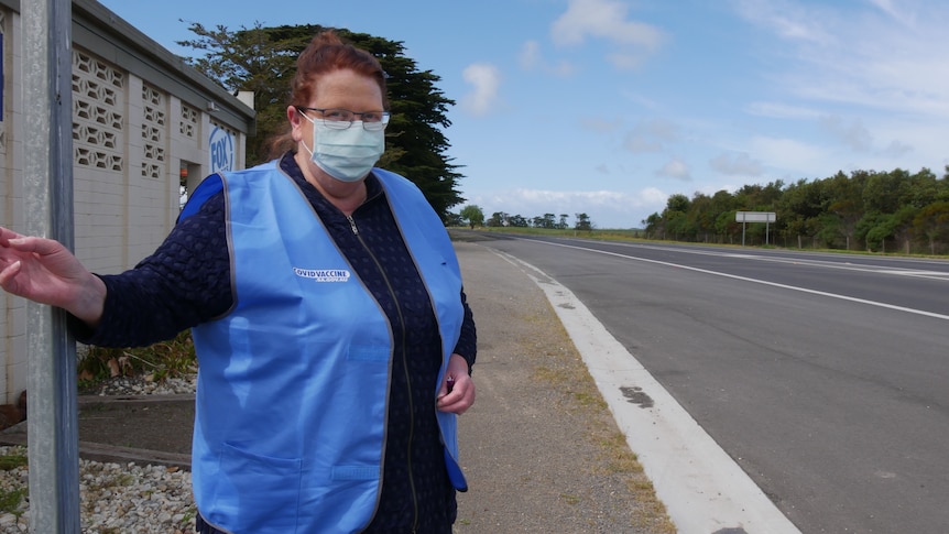 A woman in a blue vest and mask stands next to a roadway