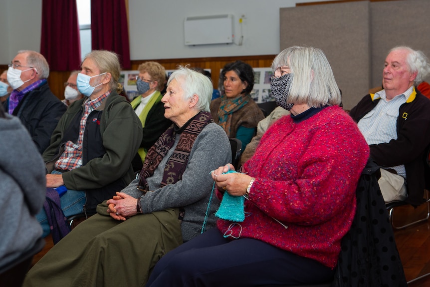 People, many with d=face masks, seated in a small town hall, looking to the stage. One lady knitting.