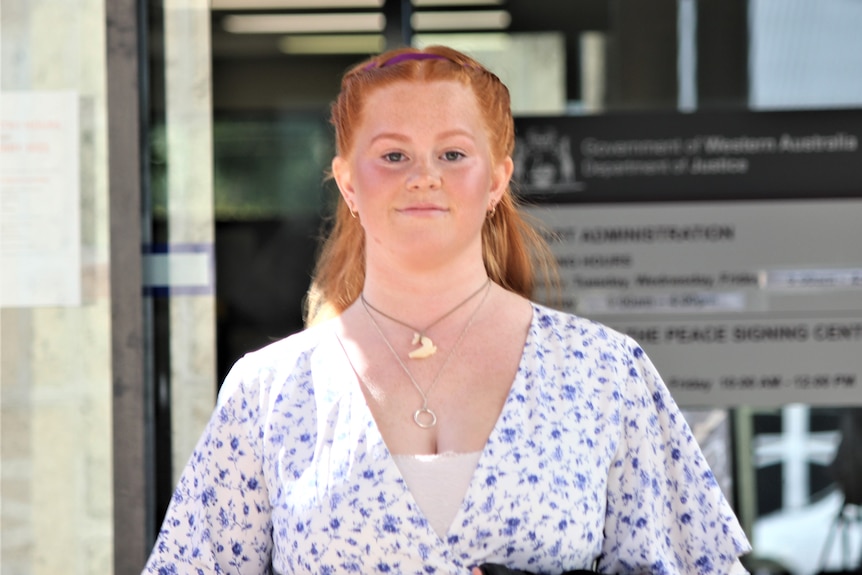 A young woman with red hair in a white dress with blue flowers, outside a court building.