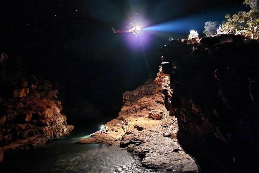 People sitting on a rock surface at night. A helicopter hovers above and shines a light down.