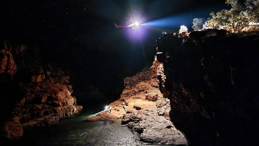 People sitting on a rock surface at night. A helicopter hovers above and shines a light down.