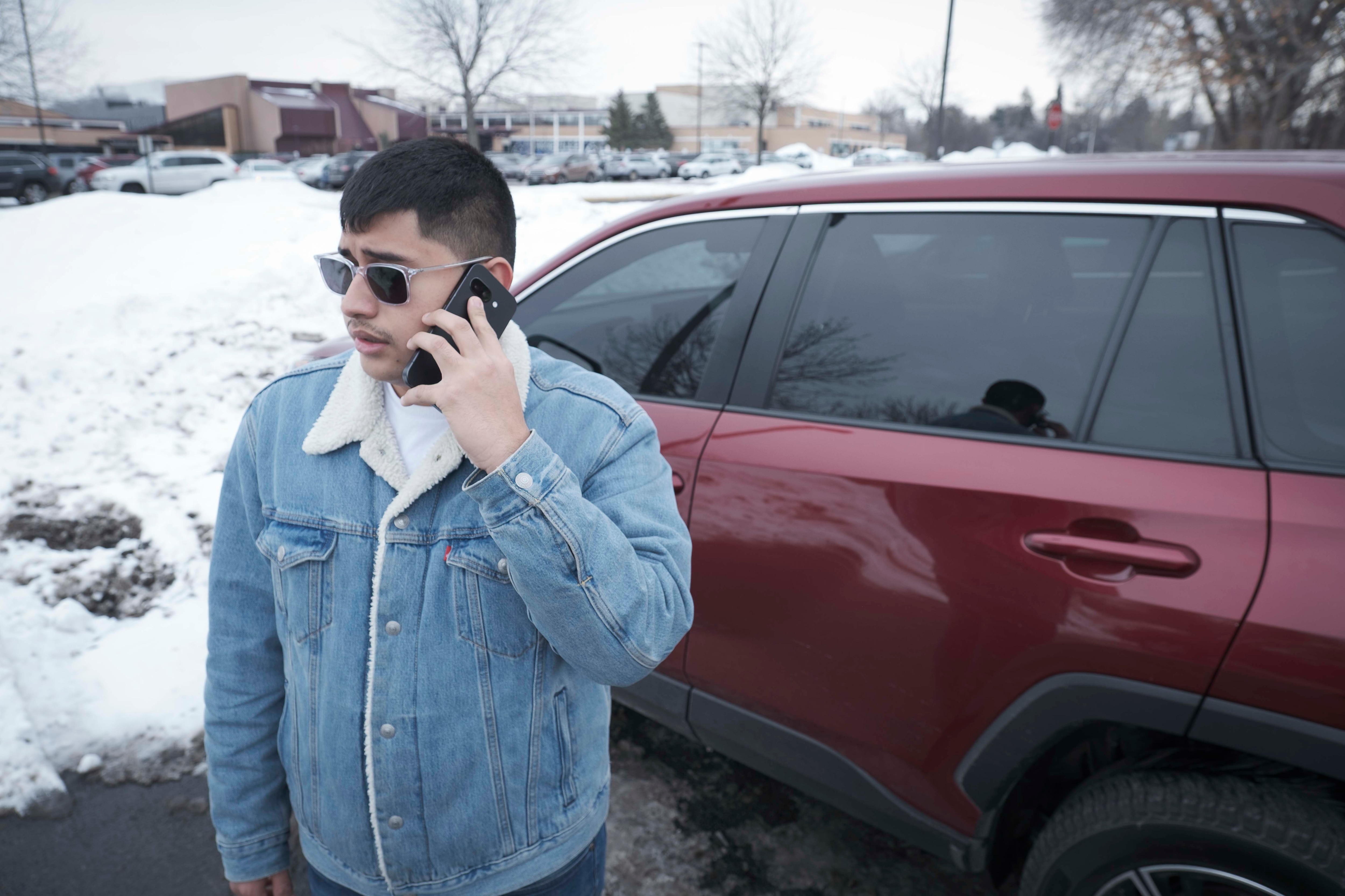 A man in a denim jacket stands next to a maroon car, speaking on the phone.