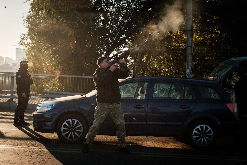 A police officer in camouflage holds a gun to the sky and shoots while standing on a road in front of a car. 