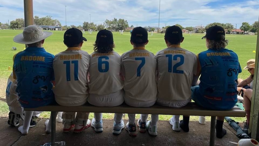 A group of young kids sit side by side on a bench, watching a cricket game take place.