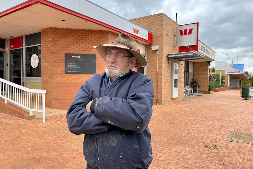 A man stands out the front of an outback bank with arms crossed wearing a hat