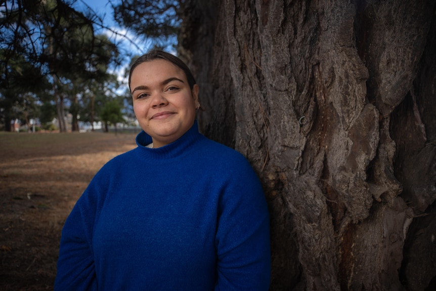 A young Indigenous woman in a blue jumper stands next to a tree and smiles