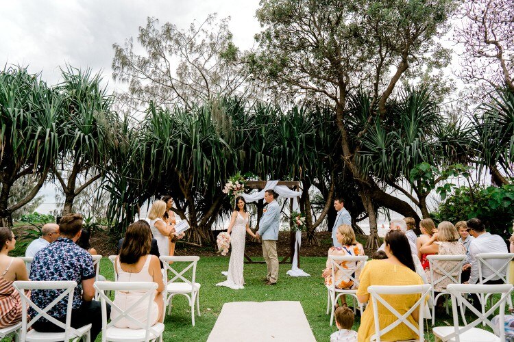 A bride and groom getting married with guests sitting on chairs and beach in background