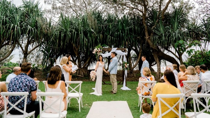 A bride and groom getting married with guests sitting on chairs and beach in background.