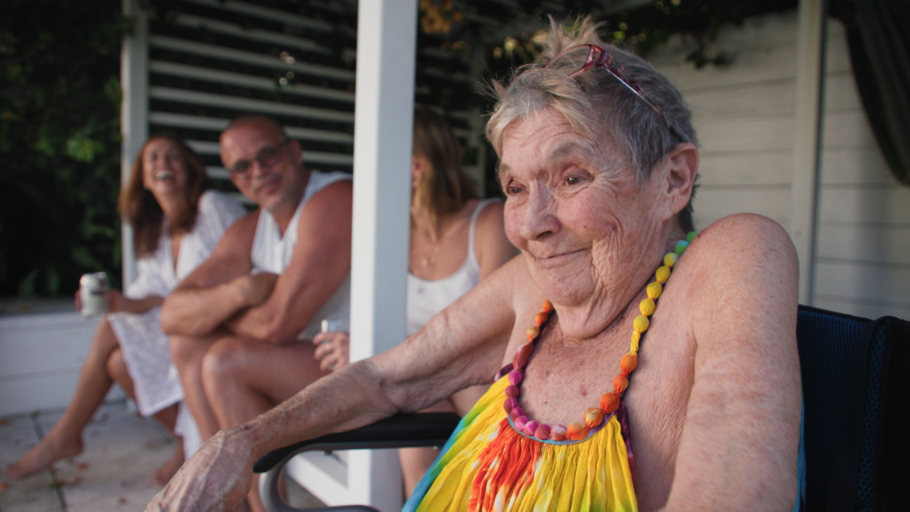 An older woman wearing a rainbow tie-dyed dress sitting in a wheelchair next to her older children who are laughing.