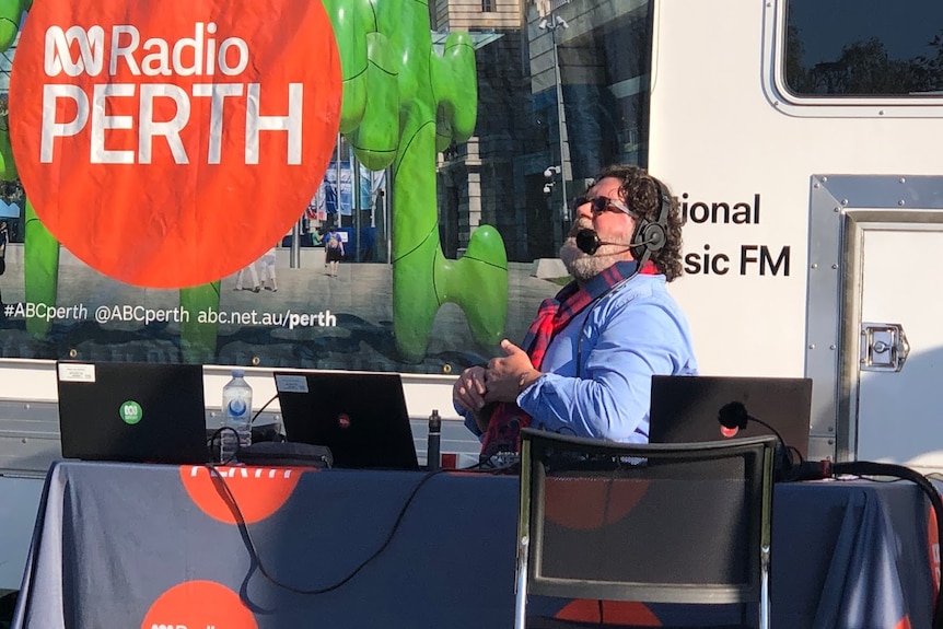 A wide shot of radio presenter Russell Woolf taking part in an outside broadcast, sitting down with headphones on.