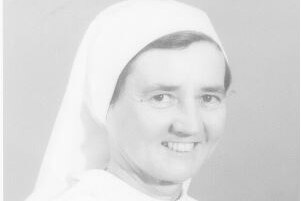 A black and white photograph of a young nun smiling. She wears white, and has her hair covered with a head piece
