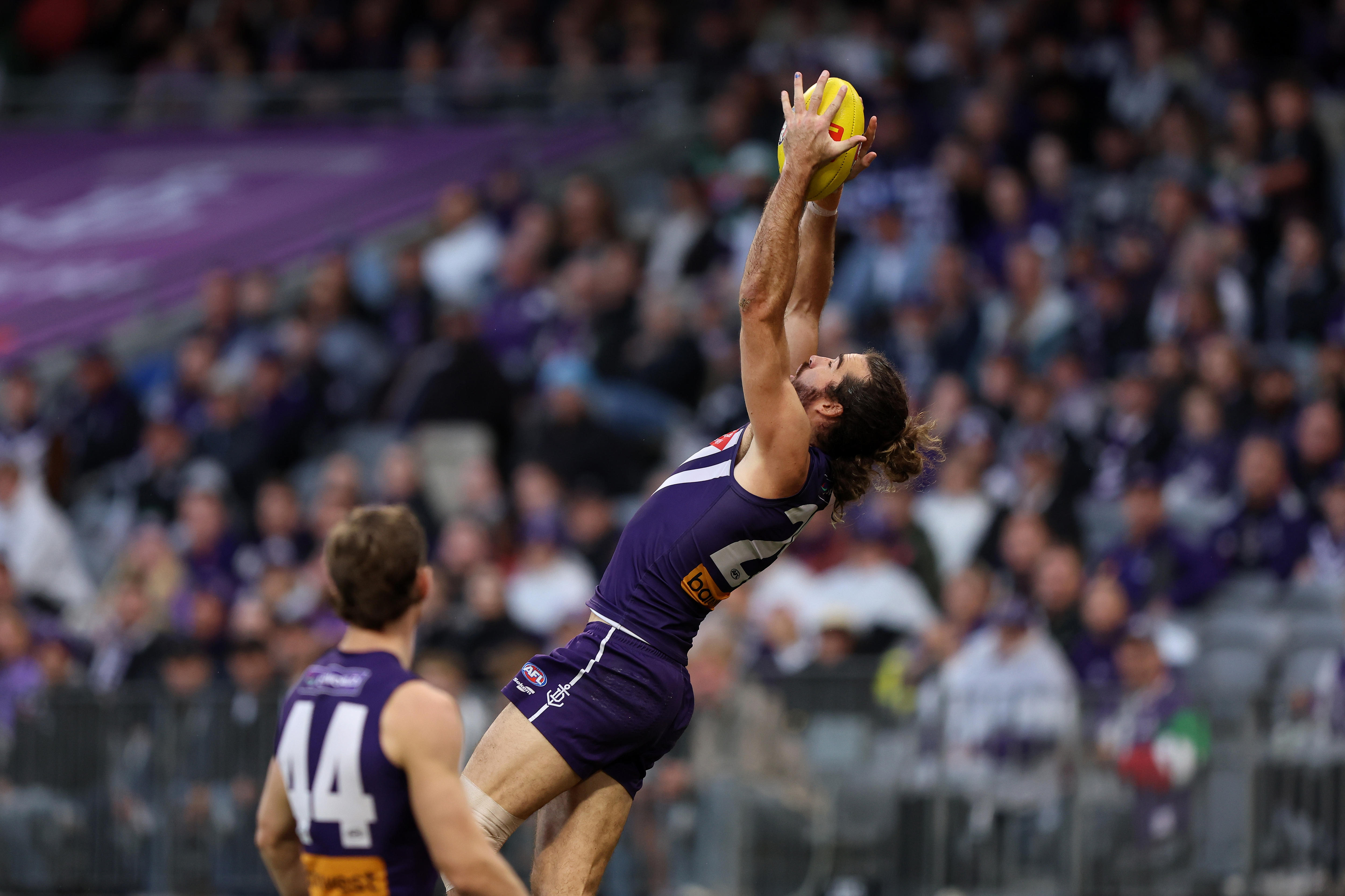 A Fremantle AFL player stretches his arms out above his head to take an athletic mark as a teammate stands by. 