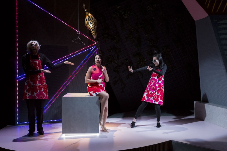 Three cast members stand on a starkly lit stage, all wearing bright pink and white floral dresses, pulling amusing faces.