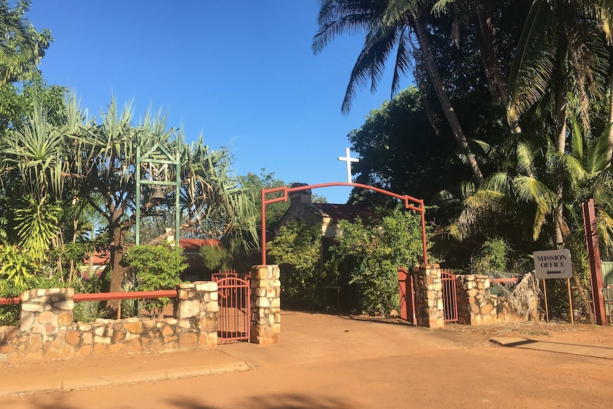 A stone wall and gate with crucifix mark surrounded by tropical vegetation and a blue sky
