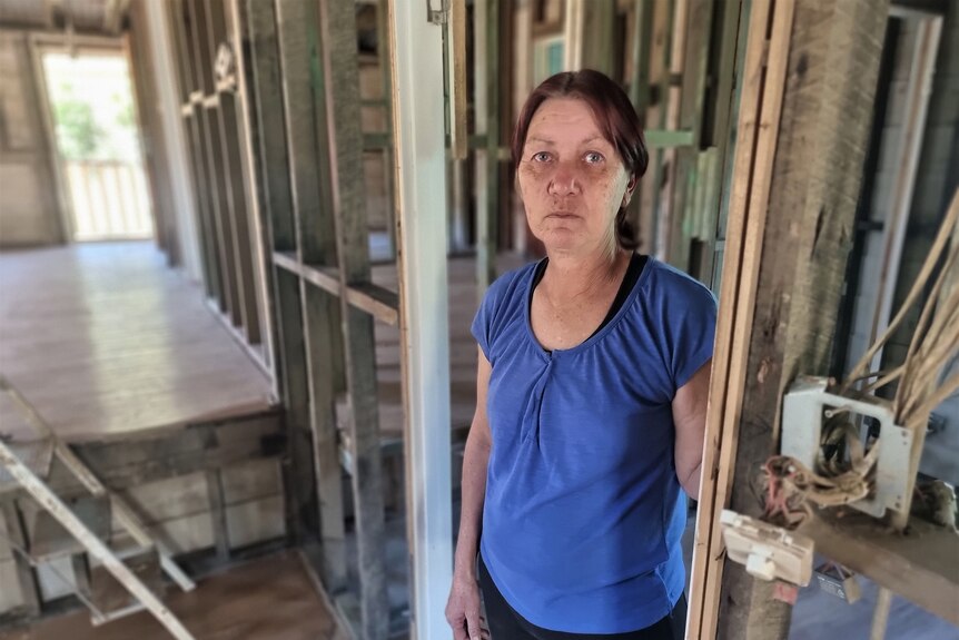 A woman with dark hair stands in the remains of a flooded home, looking glum.