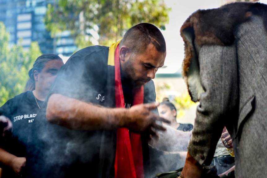 Man, wearing black top, pushes smoke onto himself in an Aboriginal smoking ceremony outside, helped by man in grey suit
