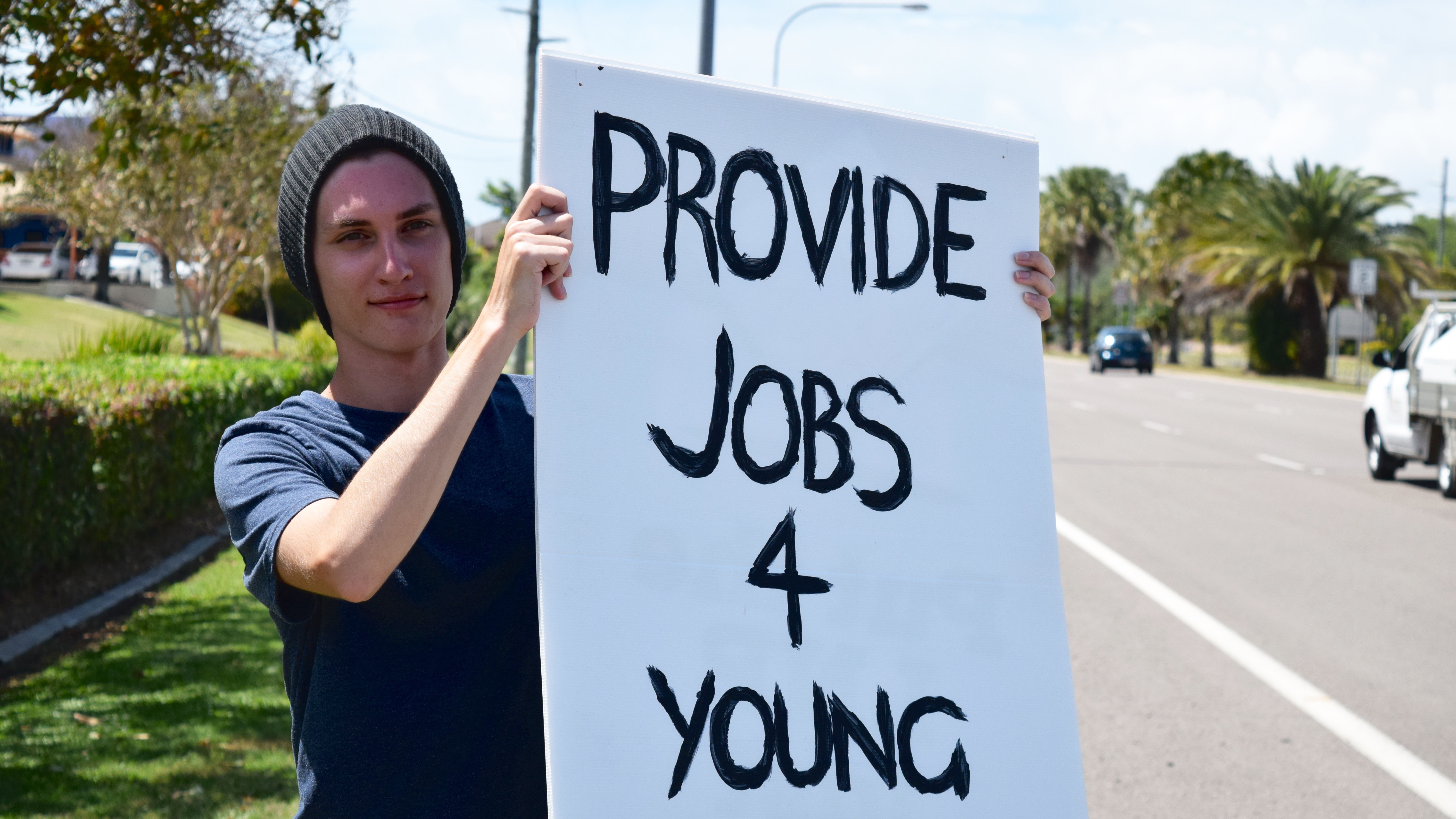 A young man wearing a beanie holds a sign that reads "provide jobs 4 young people".