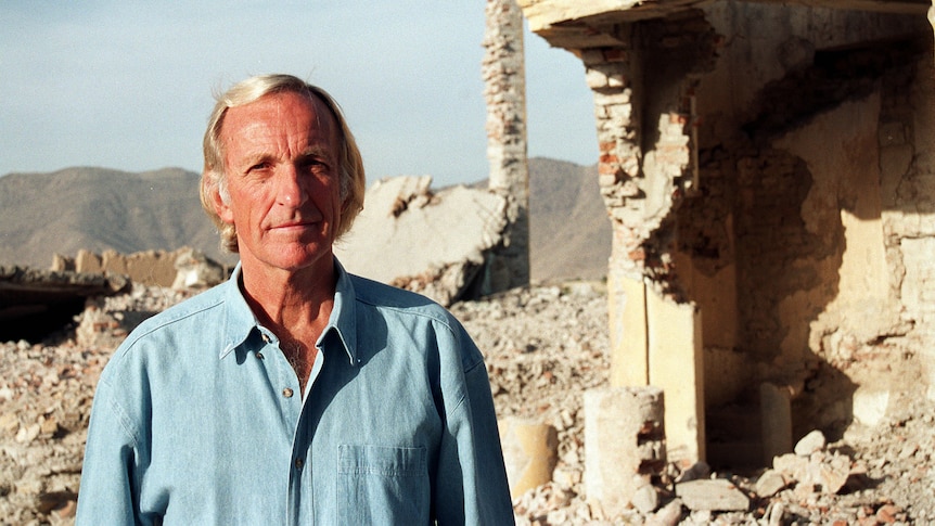 A photo of a middle-aged man standing outdoors in the sun near a damaged building.